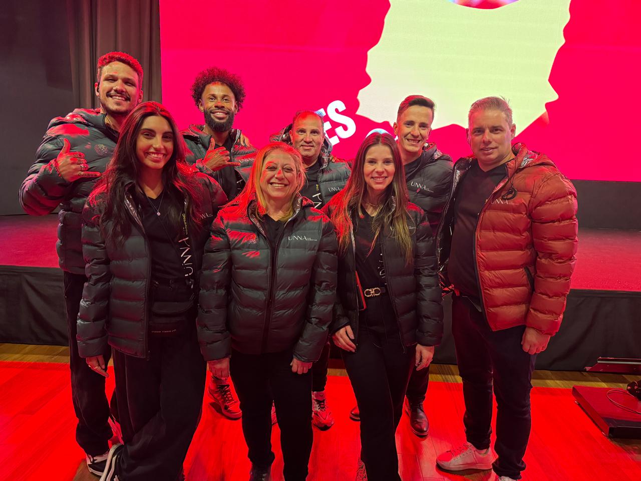 Equipe UNNA Live Marketing reunida em pé, vestindo jaquetas da marca, em evento com cenografia rosa.